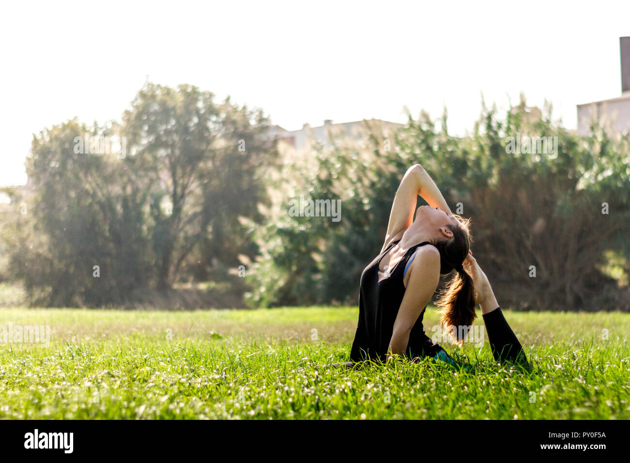 Atletica Giovane donna pratica di hatha yoga in un campo verde con erba verdeggiante accanto a un fiume su un background urbano Foto Stock