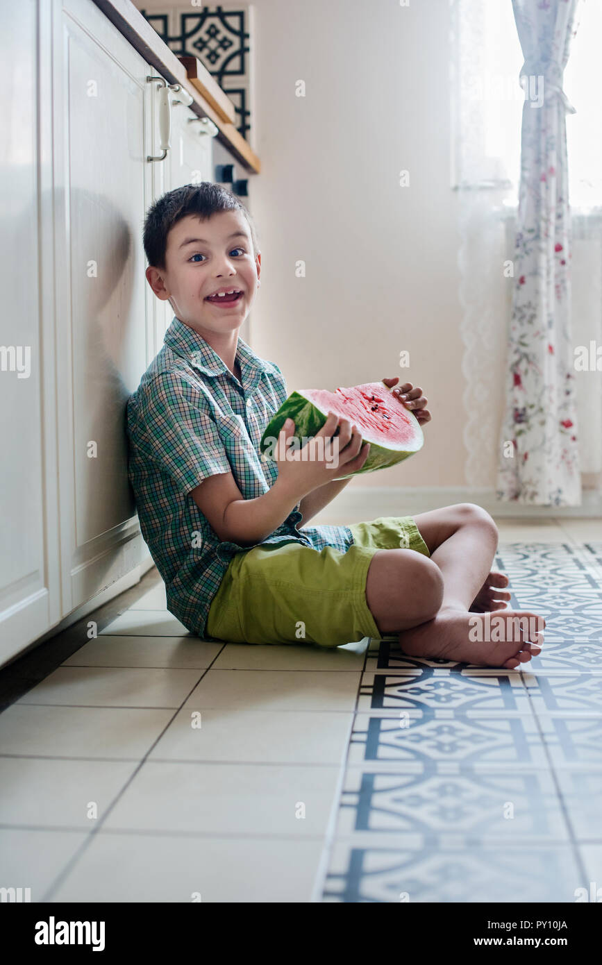 Sorridente ragazzo seduto sul pavimento della cucina tenendo una fetta di cocomero Foto Stock