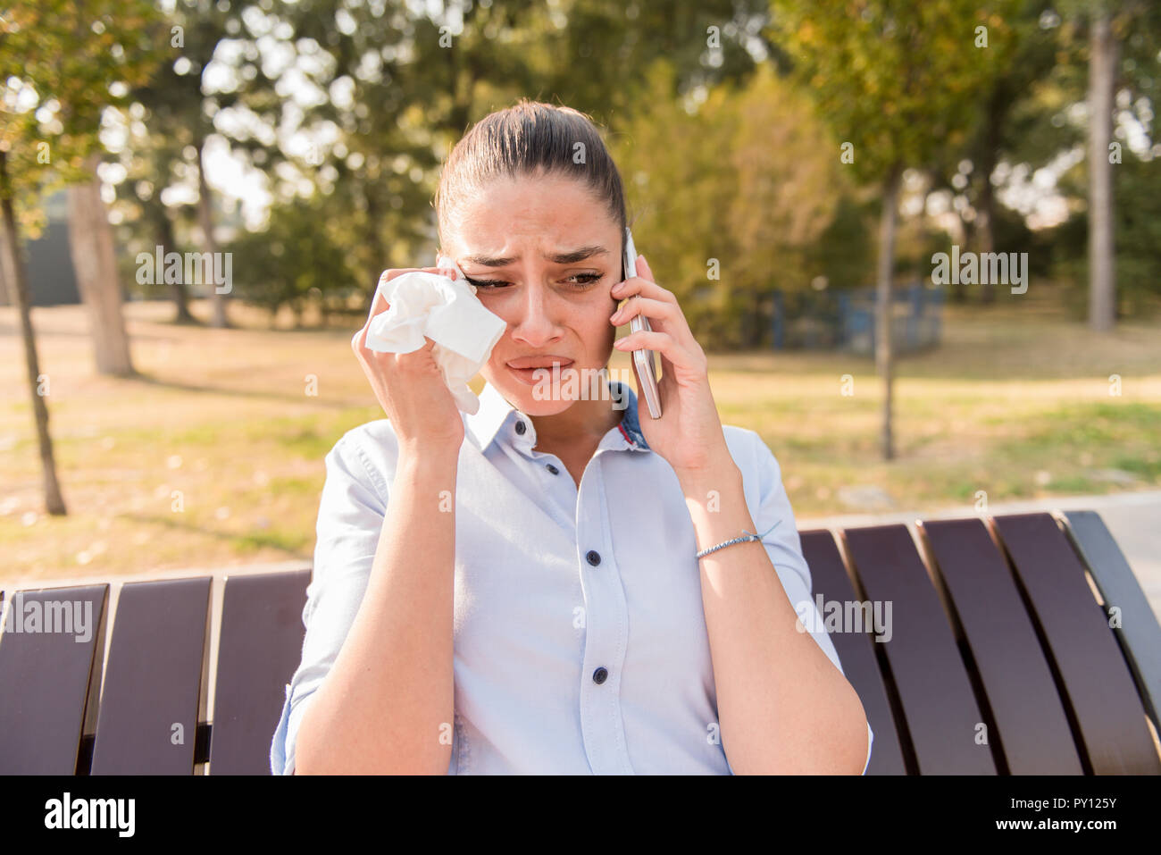 Triste giovane donna piange mentre parla al telefono mobile nel parco sul banco di lavoro Foto Stock