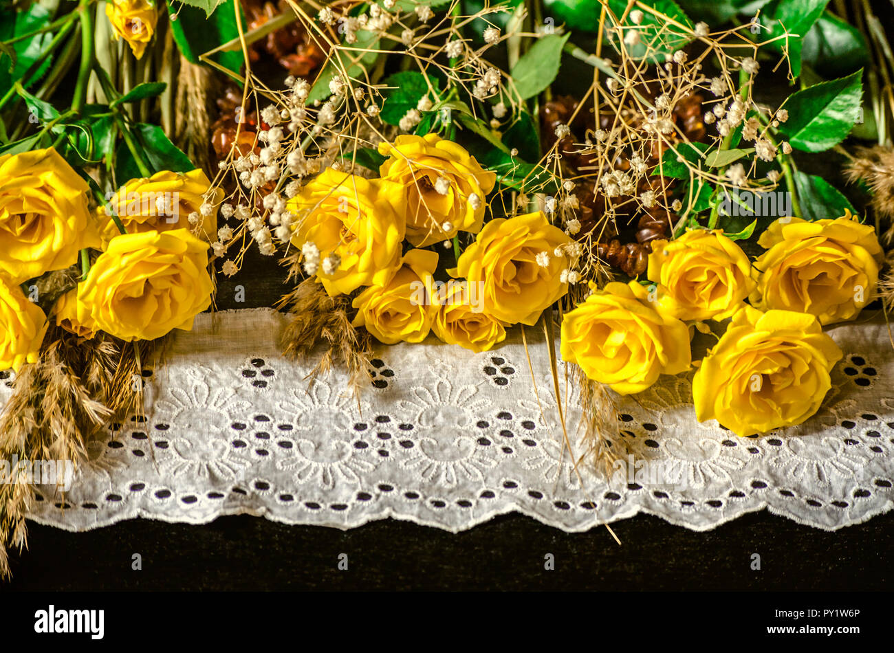 Rose di colore giallo con rami secchi gypsophila bianco e ance su bianco ricamato laici di bordatura sul legno compensato nero Foto Stock