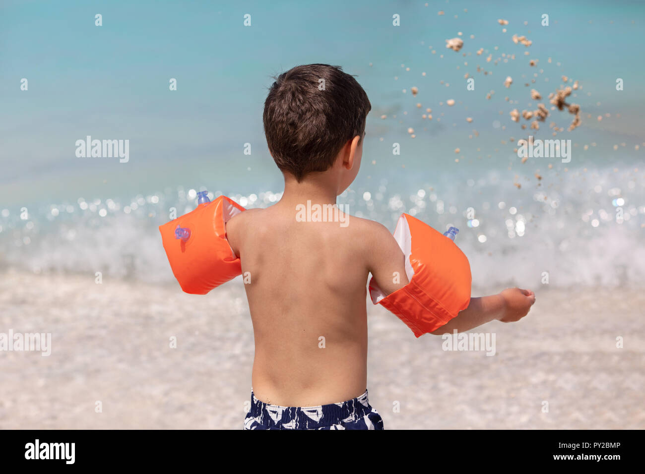 Ragazzo permanente sulla spiaggia gonfiabile indossa bracciali gettando sabbia in aria, Grecia Foto Stock