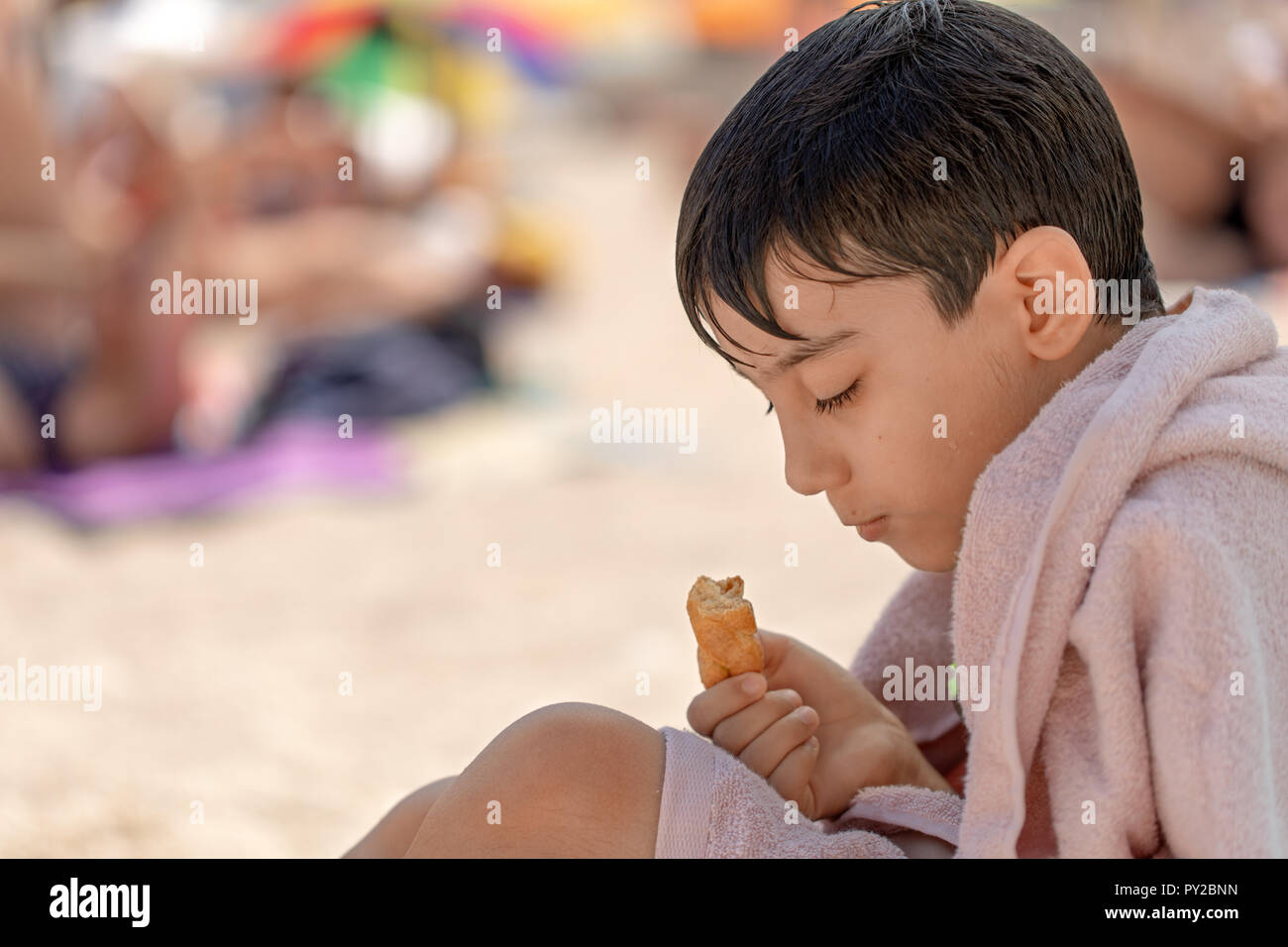 Ragazzo seduto sulla spiaggia avvolti in un asciugamano di mangiare un cornetto, Grecia Foto Stock