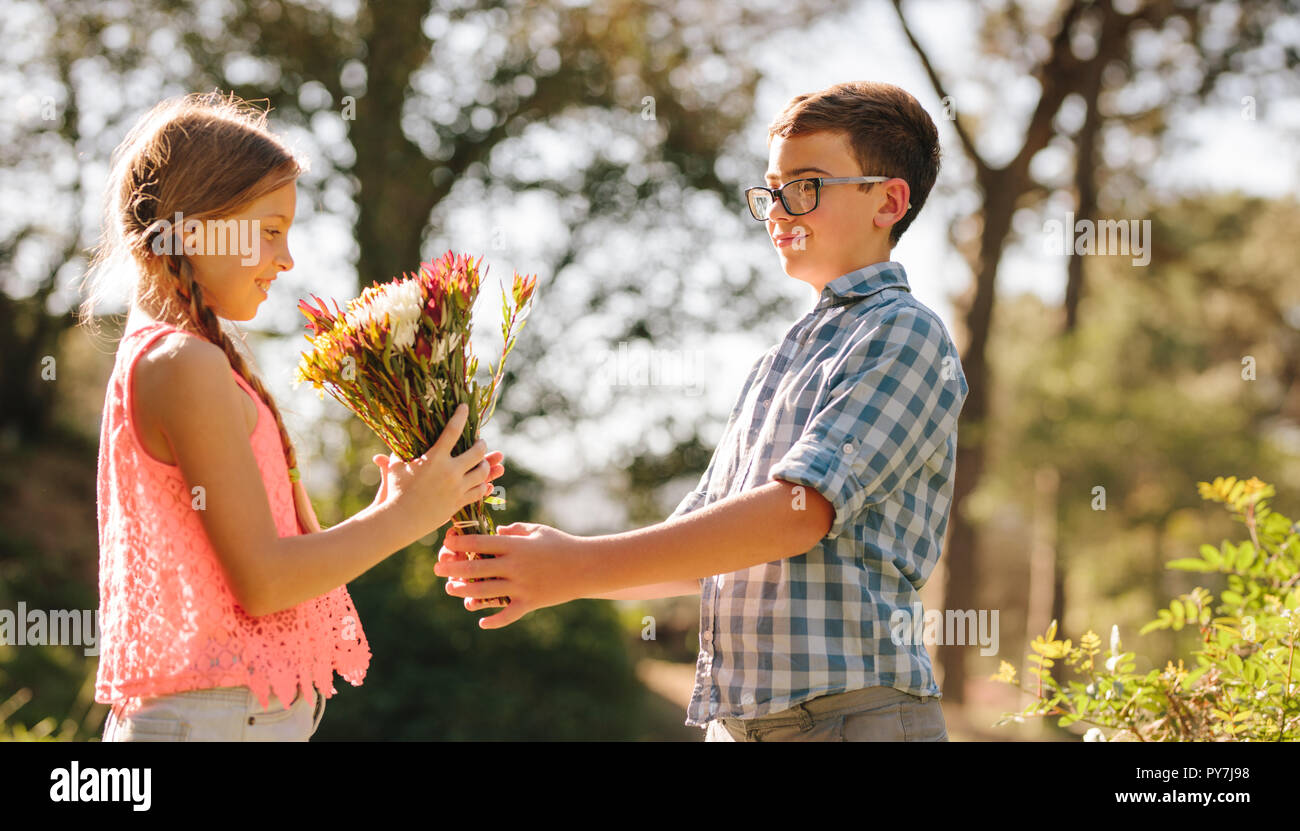 Ragazzo dando ai fiori di una ragazza in piedi in un parco. Vista laterale di amore per i bambini in piedi in un parco con un bouquet di fiori. Foto Stock