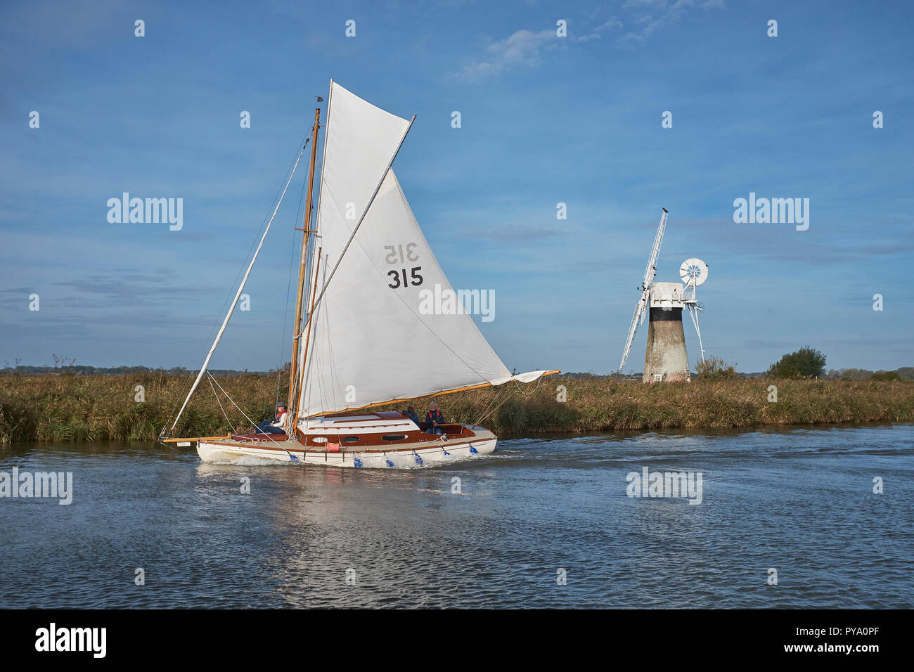 Una frazione di Sloop imbarcazione a vela passando da un mulino a vento sul fiume Thurne, su una chiara mattina autunnale, Norfolk Broads, Norfolk, Inghilterra, Regno Unito Foto Stock