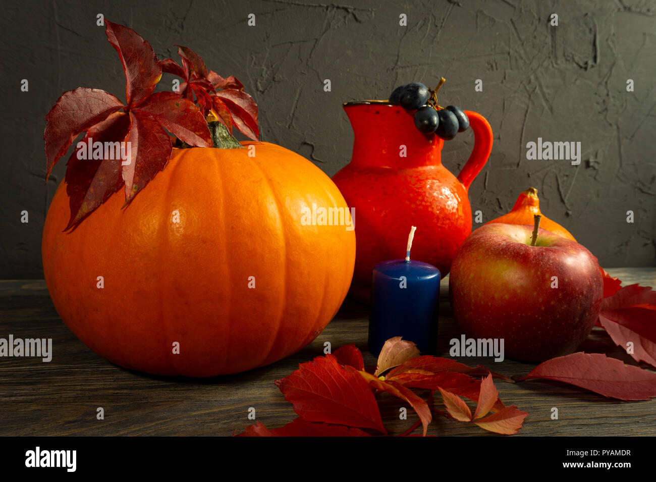 Due zucche con Bright Foglie di autunno apple, uve caraffa di rosso e blu sulla candela rustico in legno tabella contro un nero calcestruzzo ruvido grunge parete. Autunno Foto Stock