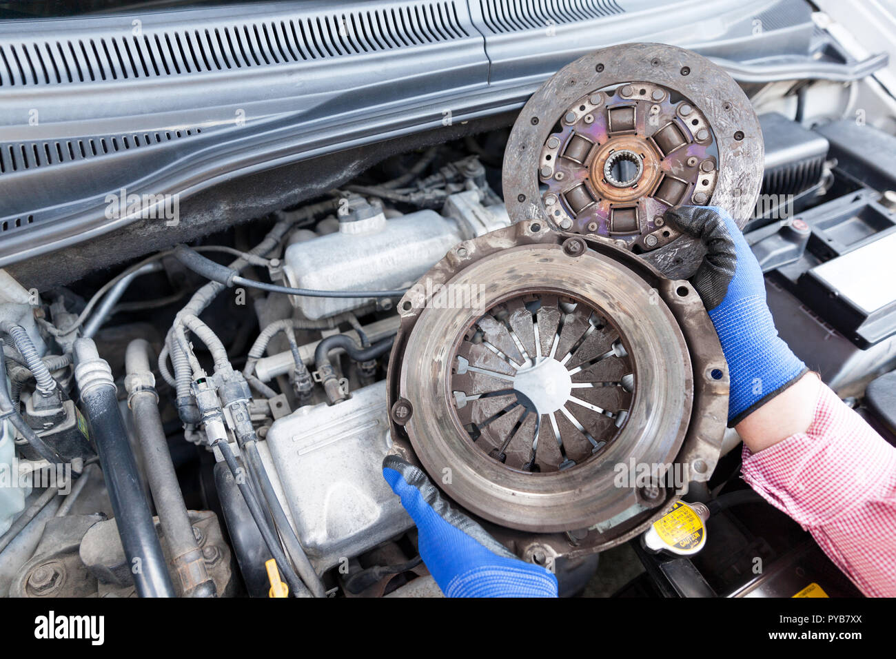 Auto meccanico di protezione da indossare guanti da lavoro tenendo un vecchio cestello della frizione e la piastra del motore sopra il motore dell'auto Foto Stock