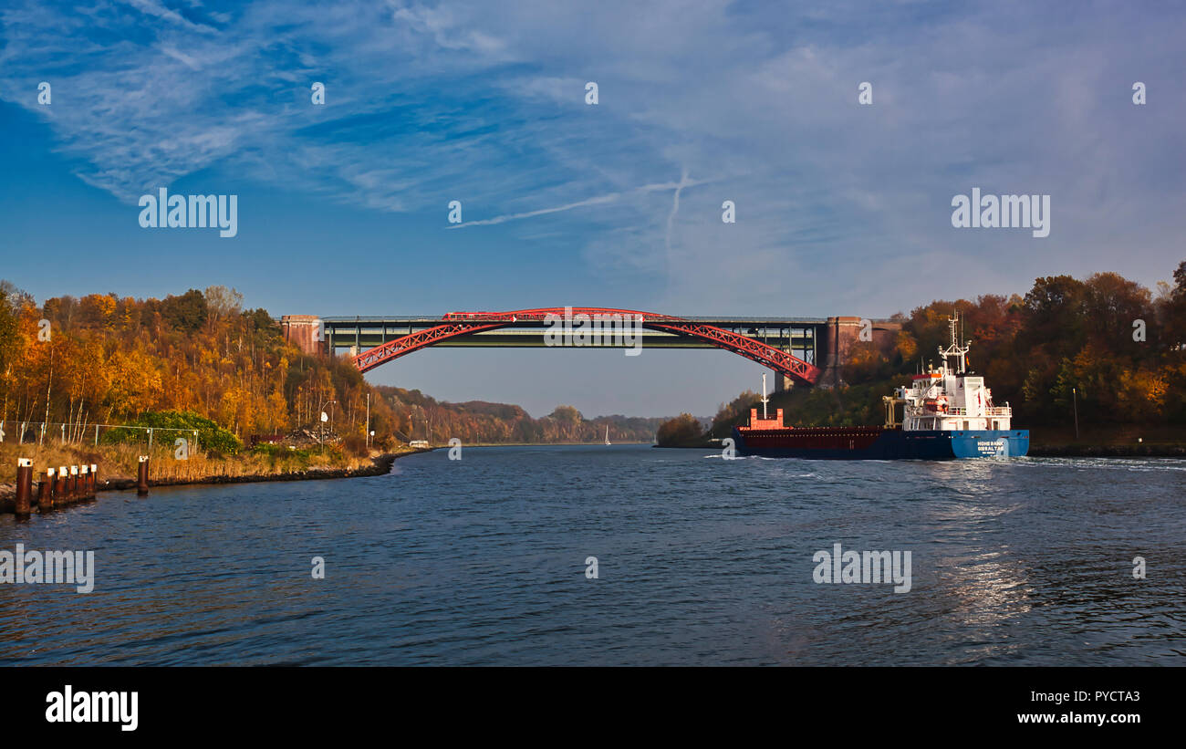 Nord-Ostsee-Kanal, 'alte Levensauer Hochbrücke', Kiel. Deutschland. Germania. Una nave portacontainer in avvicinamento al Ponte Vecchio. È tempo d'autunno e gli alberi lungo la riva del canale sono visualizzati i colori dell'autunno, ricca di rossi, marroni e gialli. Questi colori ricchi sono portati fuori dal sole che splende sulla sinistra canal bank e l'acqua è pieno di riflessioni. Un treno attraversa il ponte. Foto Stock