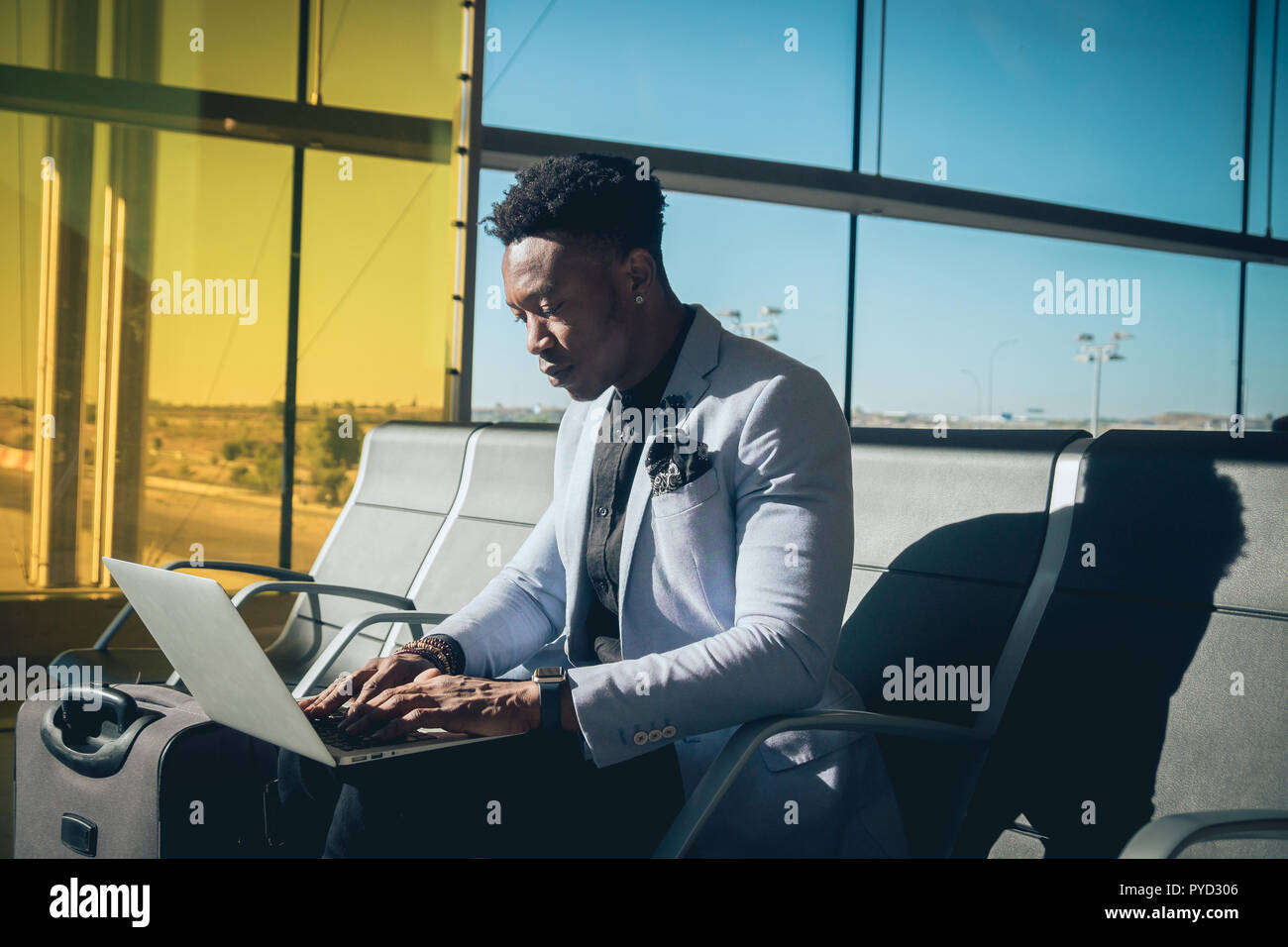 Una giovane e attraente imprenditore nero è seduto nel terminal dell'aeroporto di lavorare con un computer portatile e sorridente. Egli sta portando una valigia e indossando Foto Stock