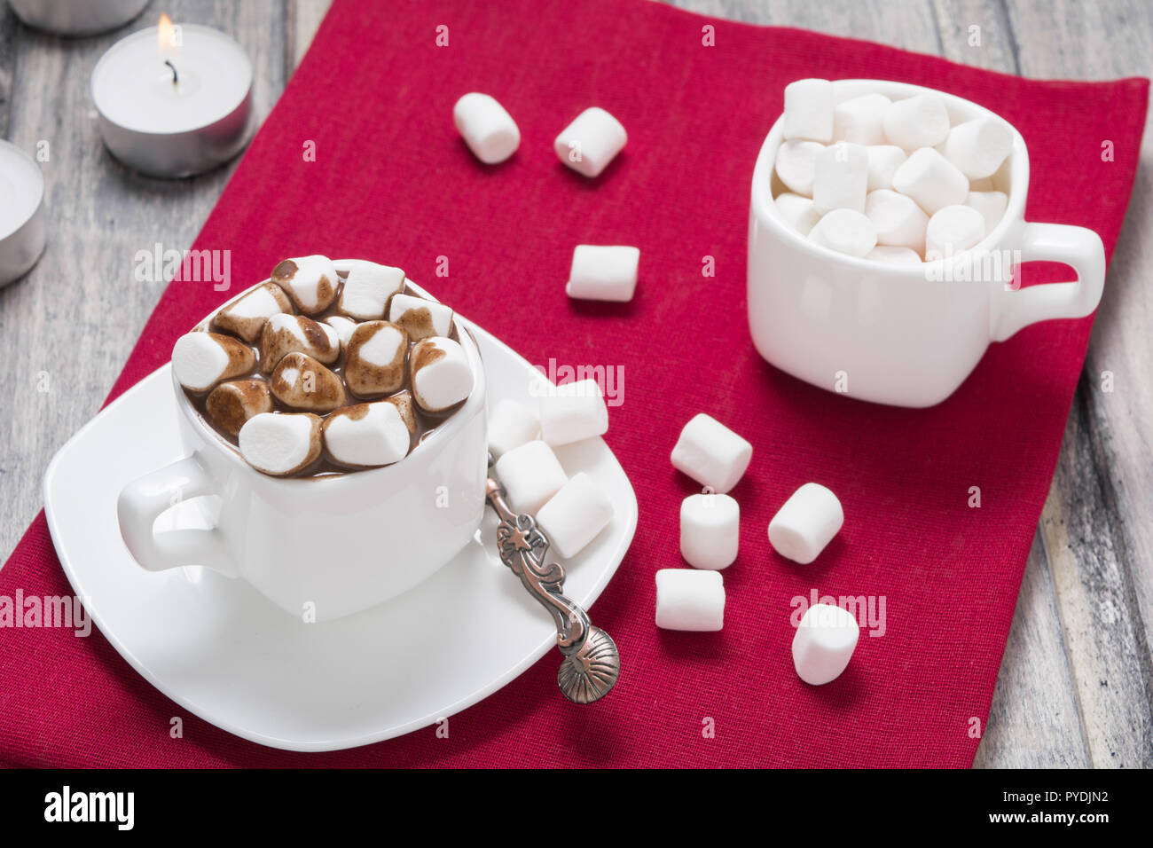 La cioccolata calda con marshmallow nella tazza bianca e due candele su asciugamano rosso sul tavolo di legno. Spazio di copia Foto Stock