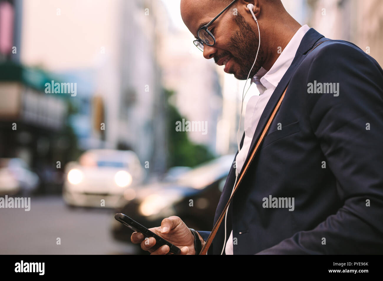 Happy businessman millenario il pendolarismo con uno smartphone. Imprenditore africano ascoltare musica attraverso le sue cuffie mentre passeggiate all'aperto sulla città Foto Stock