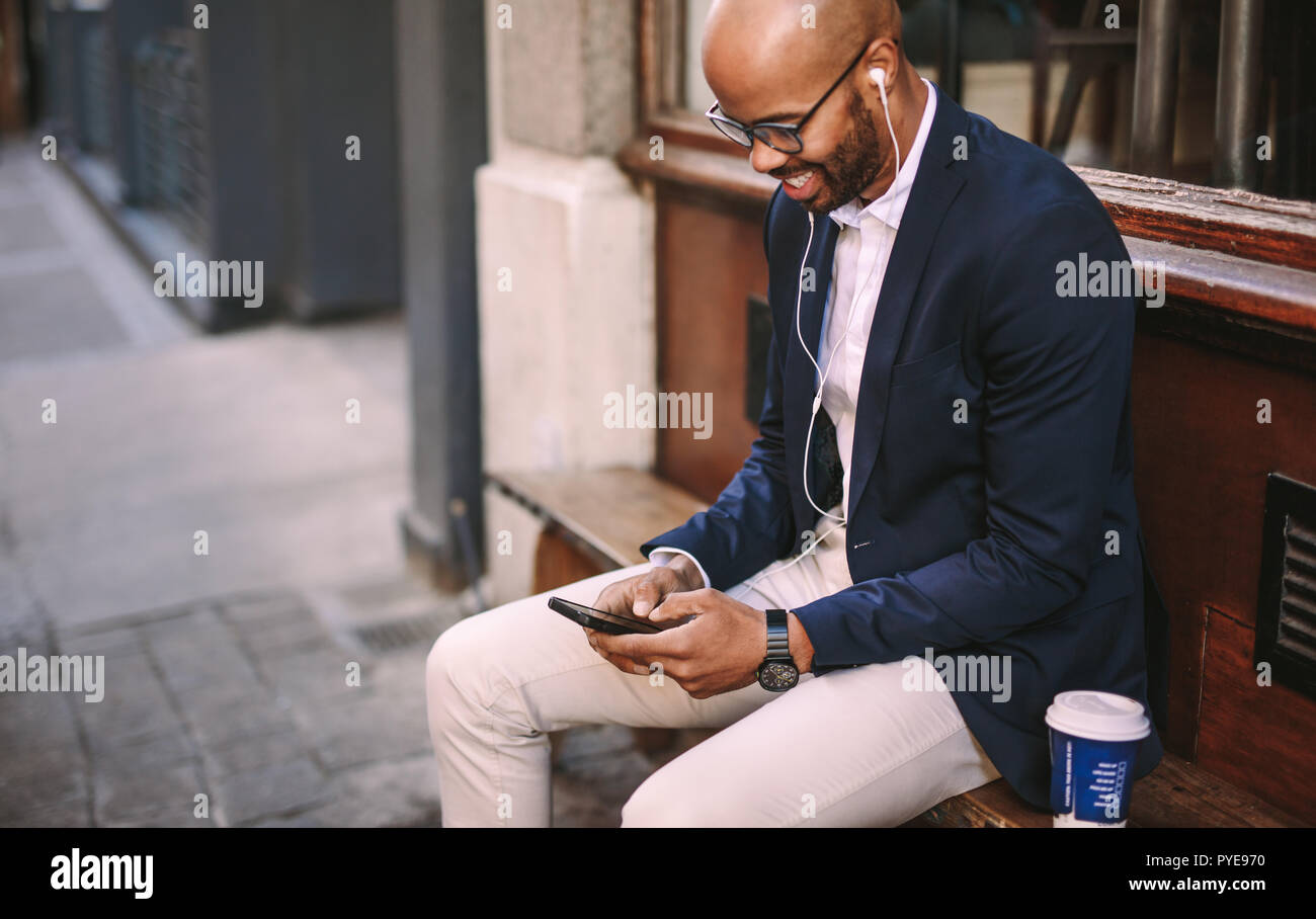 Felice giovane impreditore seduti all'aperto ascoltando la musica dal suo telefono cellulare. L'uomo africano in tuta tramite telefono cellulare al di fuori seduta. Foto Stock