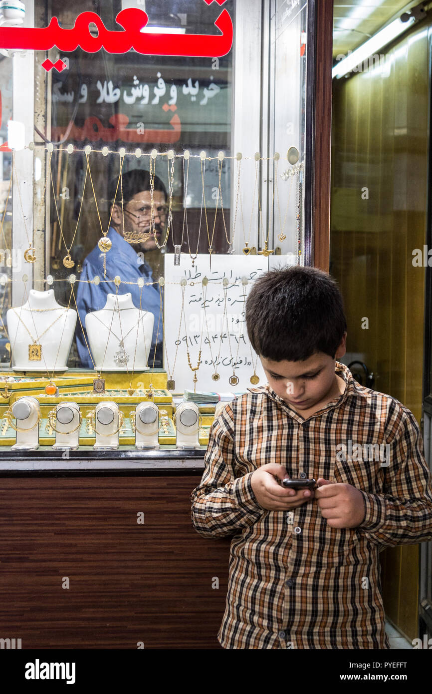 ISFAHAN, IRAN - 20 agosto 2016: Giovane ragazzo giocando con uno smartphone di fronte ad un negozio di Gioielleria in Isfahan mercato, mentre il suo vecchio padre è lookin Foto Stock