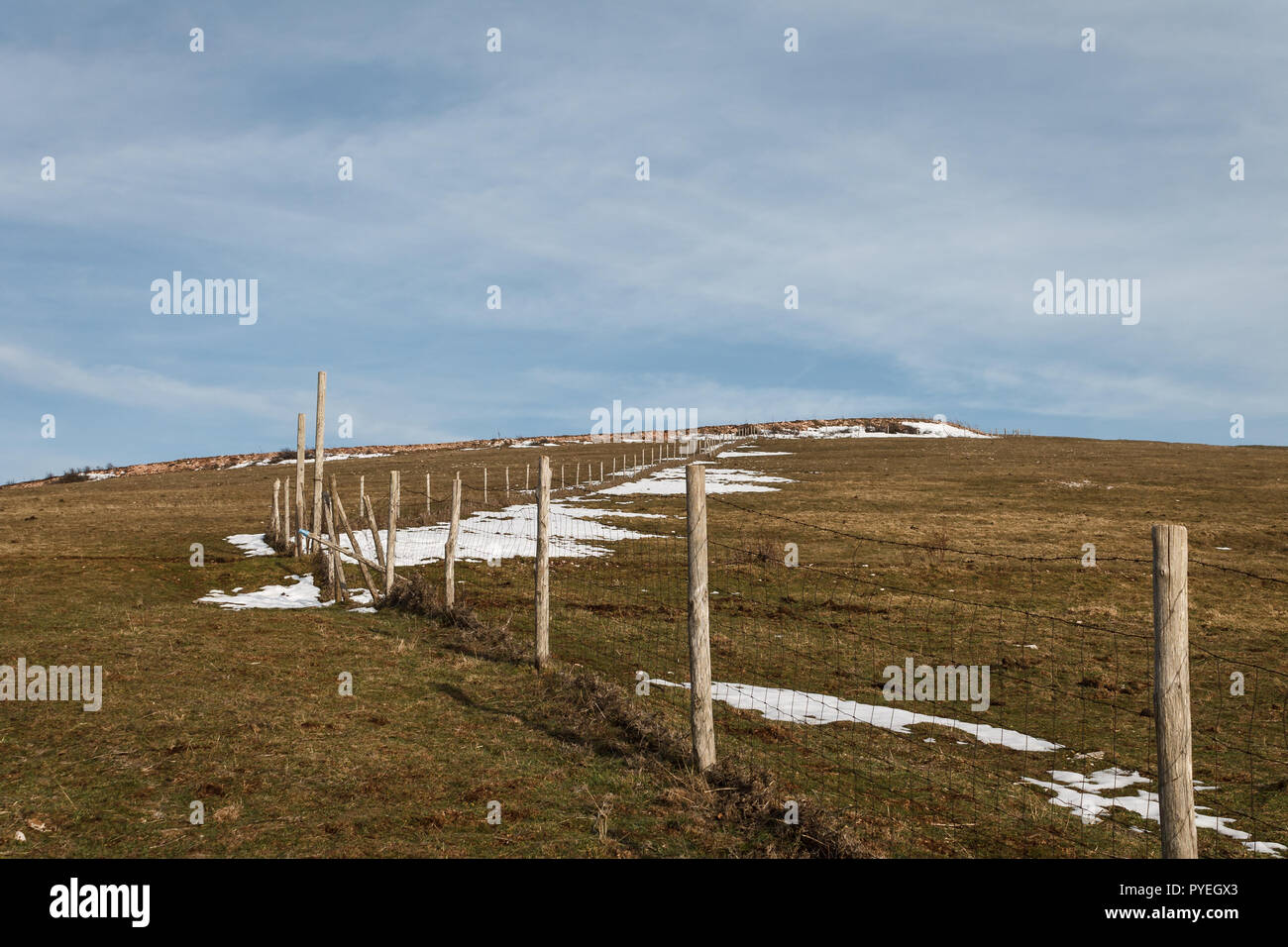 Staccionata in legno posti e filo spinato segna la linea di proprietà di una prateria con erba secca e macchie di neve sotto il cielo blu con nuvole bianche Foto Stock