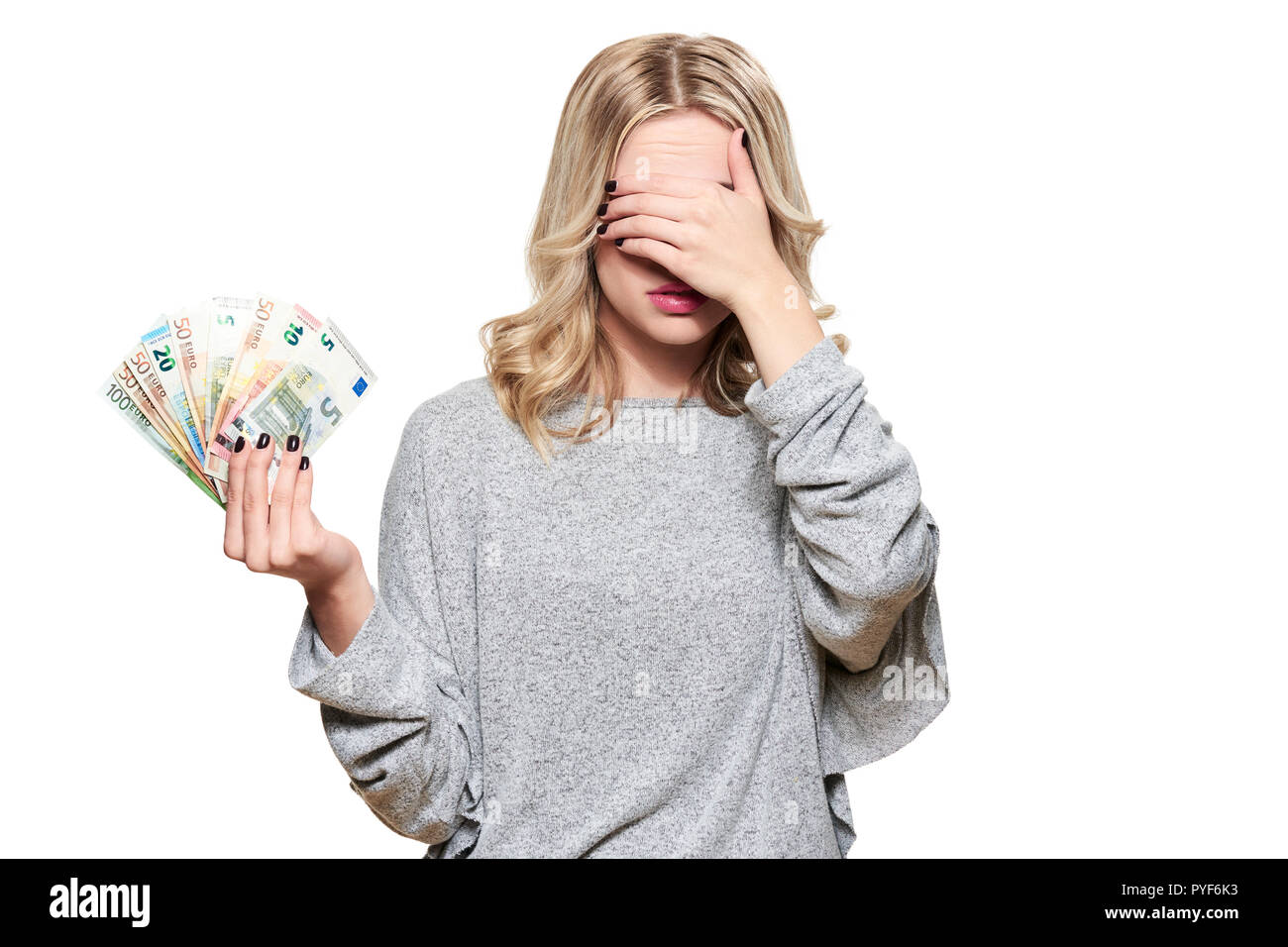 Piuttosto giovane donna in grigio maglione holding mazzetto di banconote in euro, coprendo i suoi occhi con la mano, isolato su sfondo bianco. Foto Stock