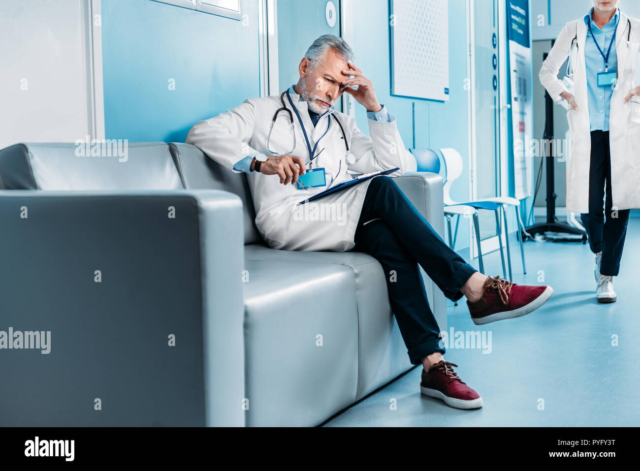 Riflessivo di mezza età medico maschio con appunti seduto sul divano mentre il suo collega di sesso femminile a piedi dietro nel corridoio di ospedale Foto Stock