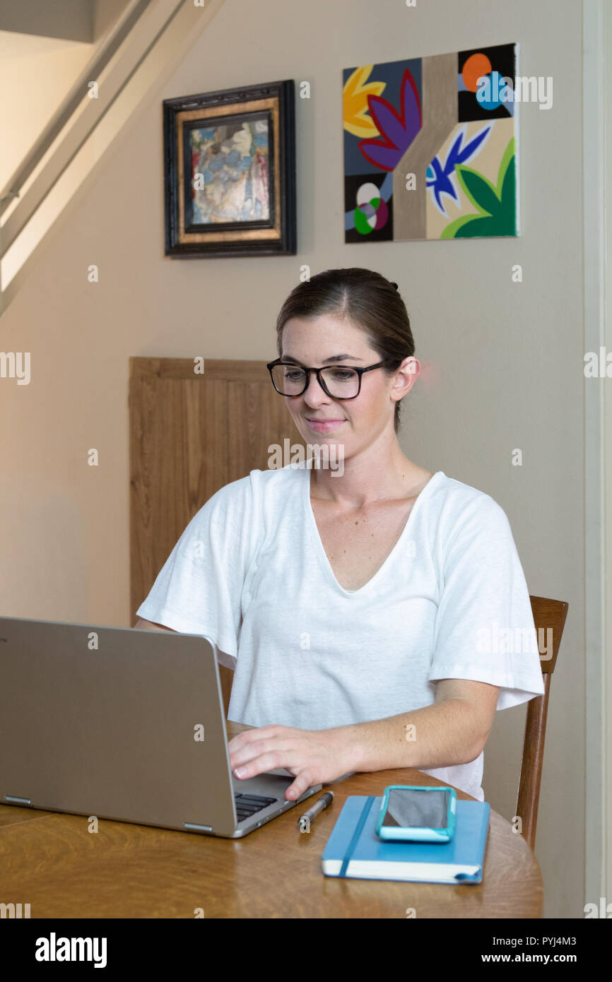 Attraente giovane donna lavora su computer presso la sala da pranzo con la sua opera/dipinto sulla parete dietro e dei dispositivi tecnologici. Foto Stock