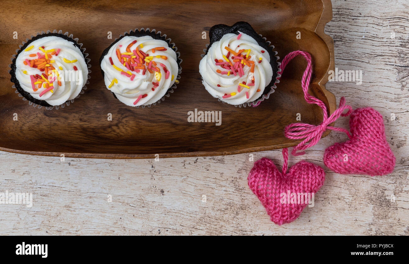 3 tortini di cioccolato con glassa alla vaniglia e spruzza su un vassoio di legno con maglia rosa cuori su una luce sullo sfondo di legno Foto Stock