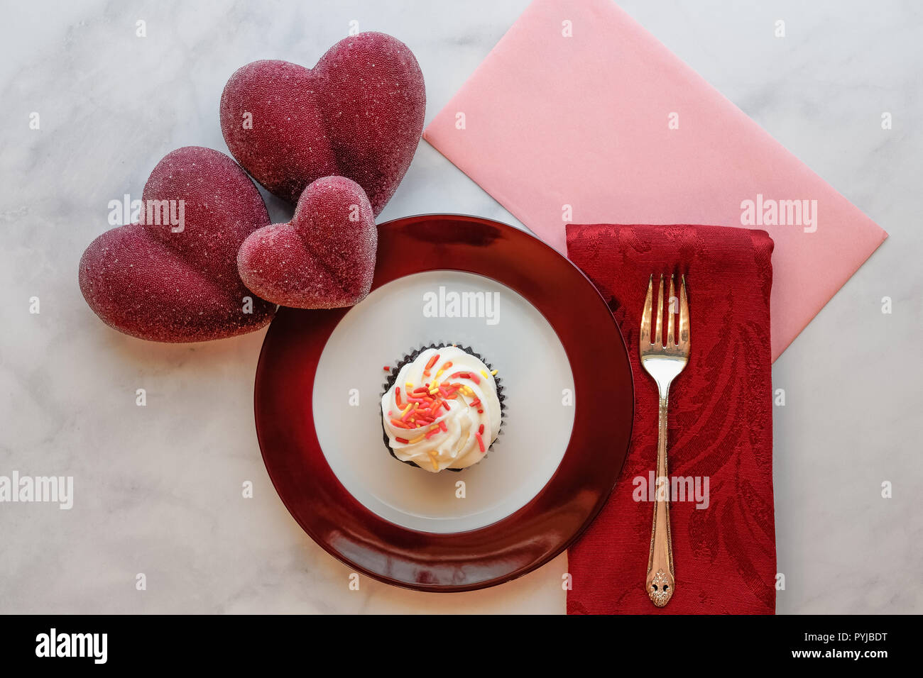 Valentines Day Treat per qualcuno di speciale con cupcake sul rosso piastra bordata, rosa biglietto di auguri busta, igienico e la forcella nonché cuore rosso ornamenti Foto Stock