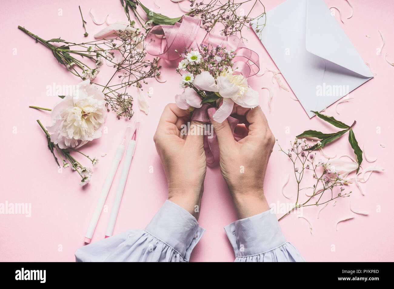 Mani femminili rendendo bellissimi fiori disposizione con le peonie fiori sul rosa pastello sfondo con busta, vista dall'alto Foto Stock