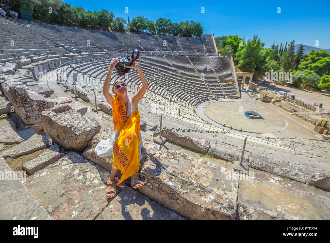 Fotografo di viaggio prendendo selfie con fotocamera reflex. Donna seduta sui gradini del Teatro di Epidaurus, prende le immagini nel sito archeologico, Peloponneso e Grecia. Vacanze estive lifestyle. Foto Stock