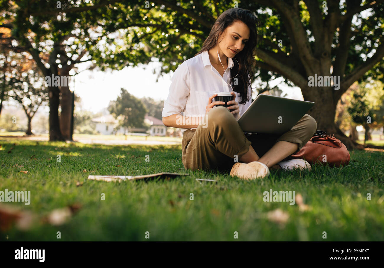 Bella donna seduta sul prato presso il park tenendo un caffè e guardando il computer portatile. Giovane imprenditrice in abiti casual seduti al park w Foto Stock