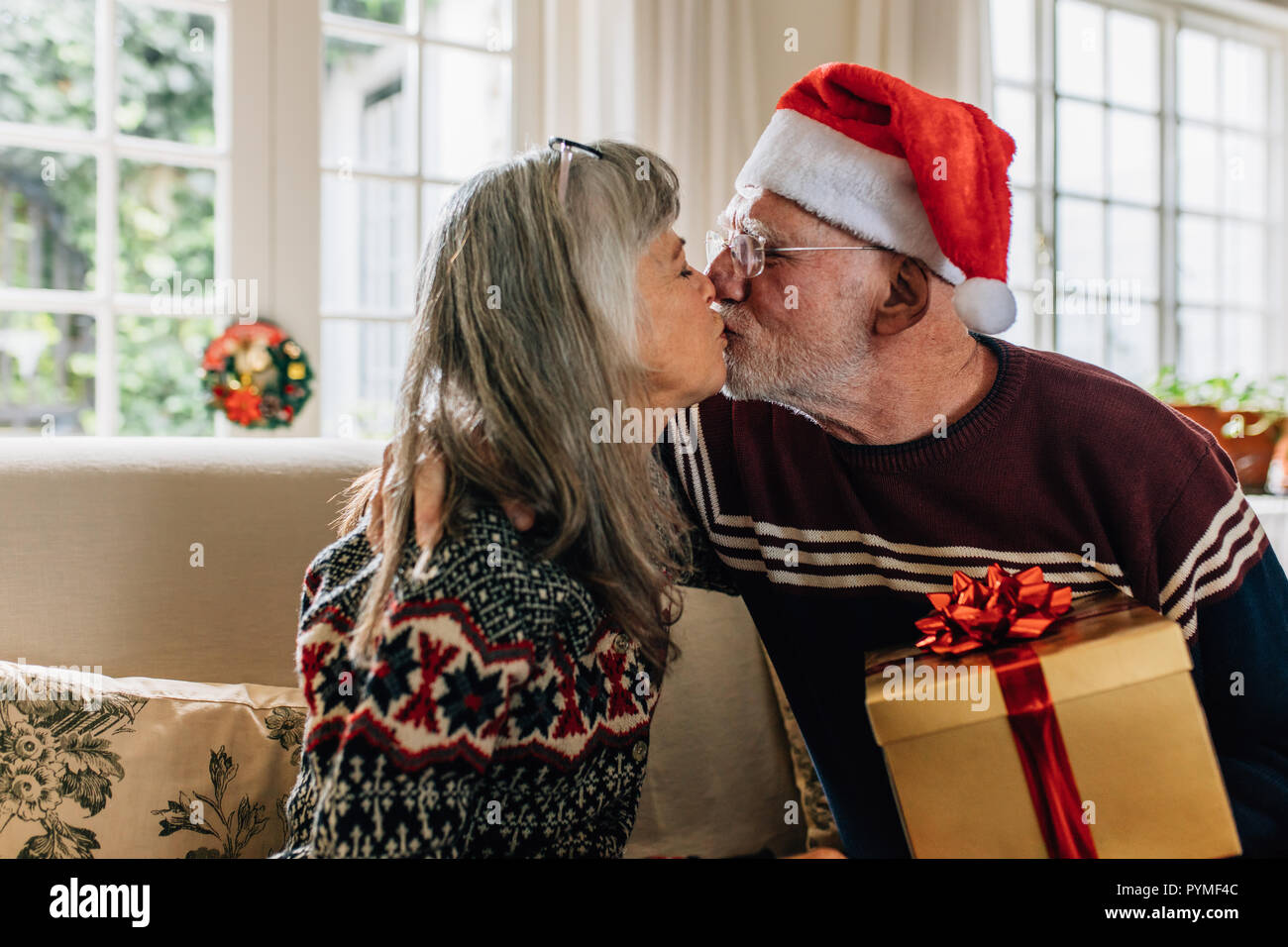 Senior uomo in santa cap baciando la moglie tenendo un regalo di Natale. Coppia senior festeggiano il Natale e il messaggio di saluto di ogni altro a casa. Foto Stock