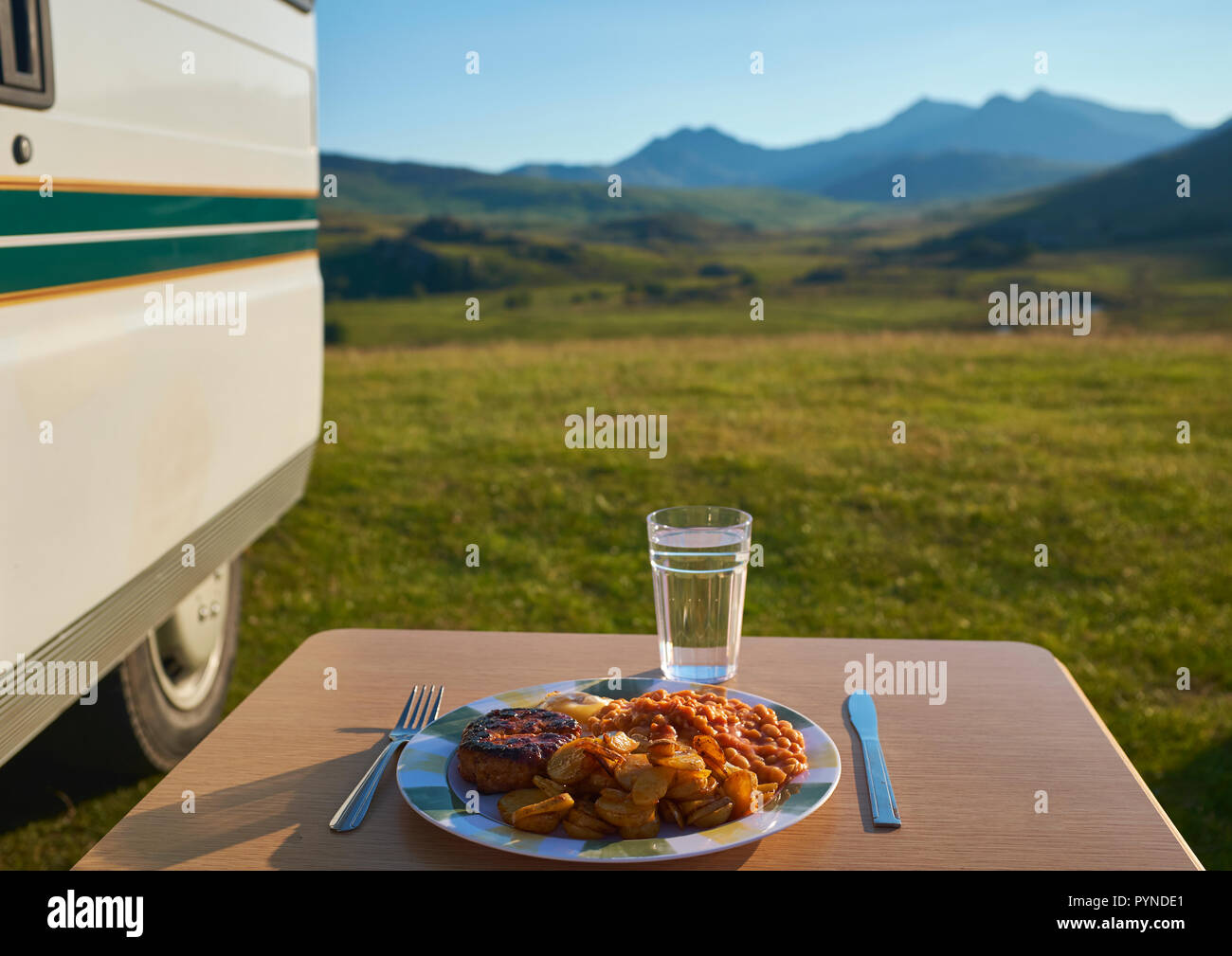 Un piatto di cibo su una tabella con la lama di un coltello, forchetta e un bicchiere di acqua all'esterno accanto a un camper con snowdon nel retro terra, Wales, Regno Unito Foto Stock
