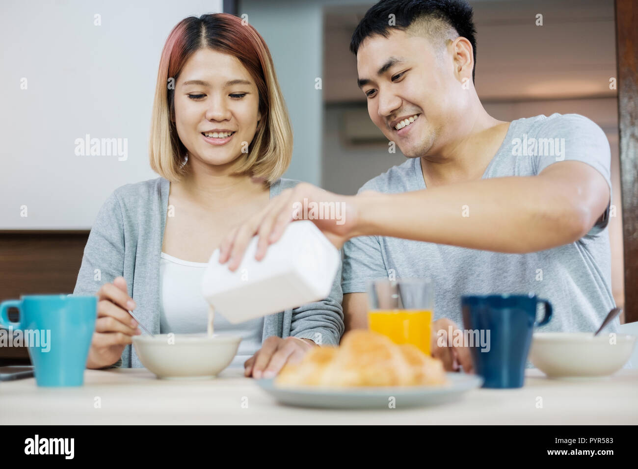 Felice coppia asiatica in pigiama seduta a tavola in cucina a casa la mattina e avente cereale colazione insieme.marito versare il latte nella tazza moglie. Foto Stock