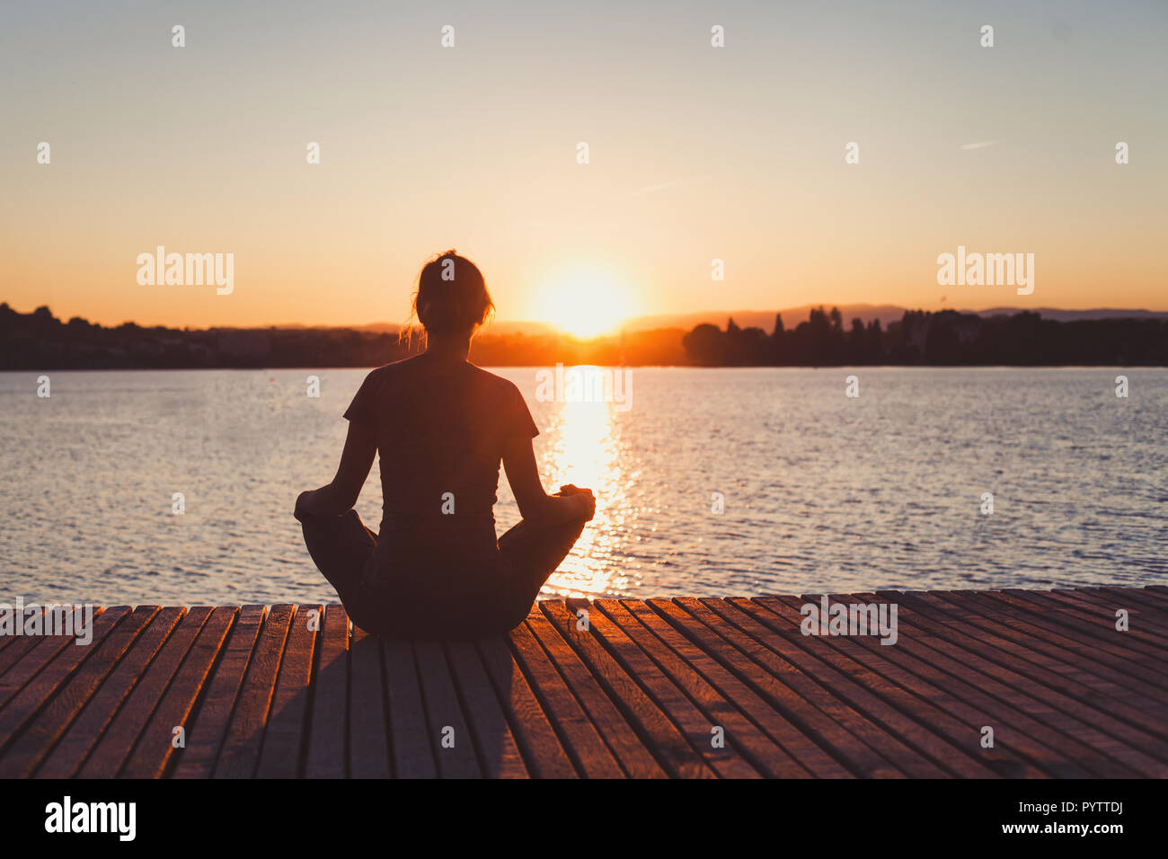 Lo yoga, donna facendo di meditazione e di esercizi di respirazione sul molo in legno vicino al lago, silhouette al tramonto Foto Stock