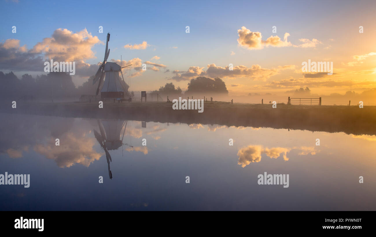 White storico mulino nella nebbia mattutina si riflette nel fiume sotto la bellissima alba, Drenthe, Paesi Bassi Foto Stock
