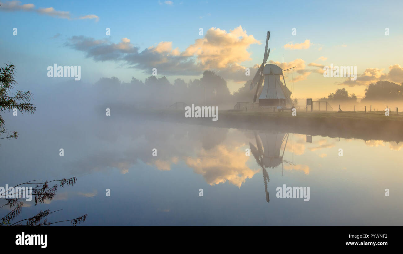 La riflessione di bianco il mulino a vento di legno nel fiume con nebbia di mattina durante il Sunrise, Drenthe, Paesi Bassi Foto Stock