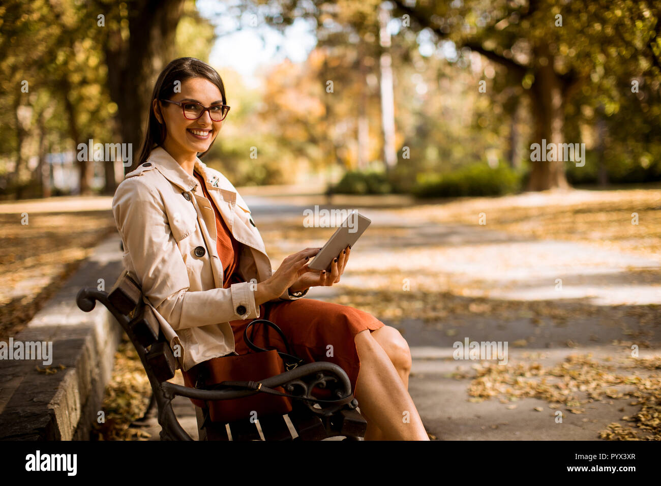 Donna seduta su un banco di lavoro nel parco durante l'autunno Meteo utilizzando un tablet PC e controllo dei mezzi di comunicazione sociale. Foto Stock
