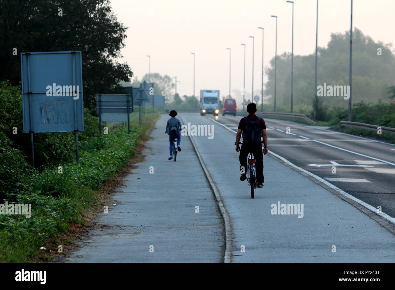 L uomo e la donna in bicicletta senza usare le mani sul marciapiede accanto alla strada con il traffico in entrata con diversi cartelli stradali e densa di erba non tagliata Foto Stock