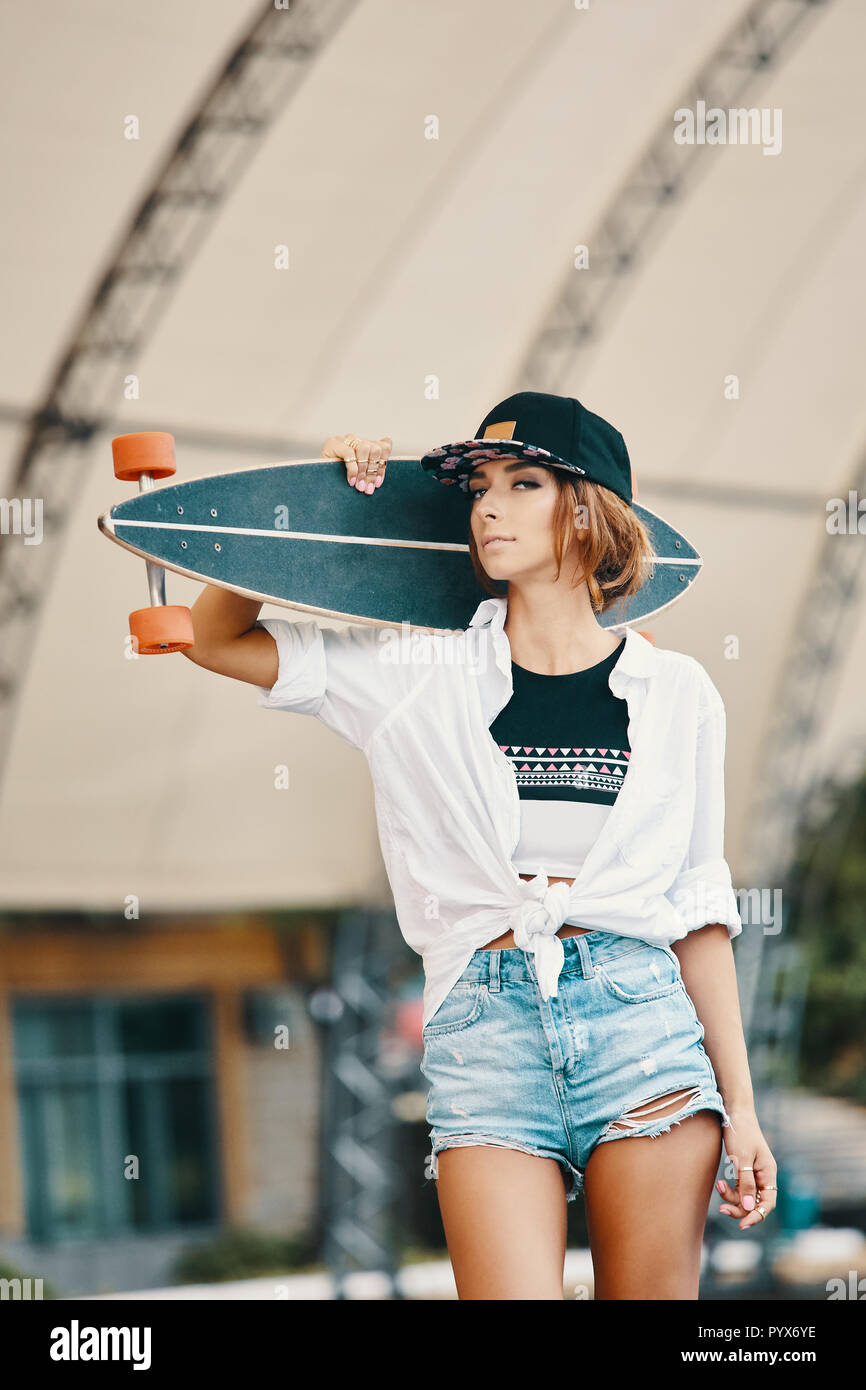 Elegante ragazza mostra atteggiamento close up ritratto, tenendo longboard sulle spalle. Sport estremo, moda giovane e concetto di emozioni Foto Stock