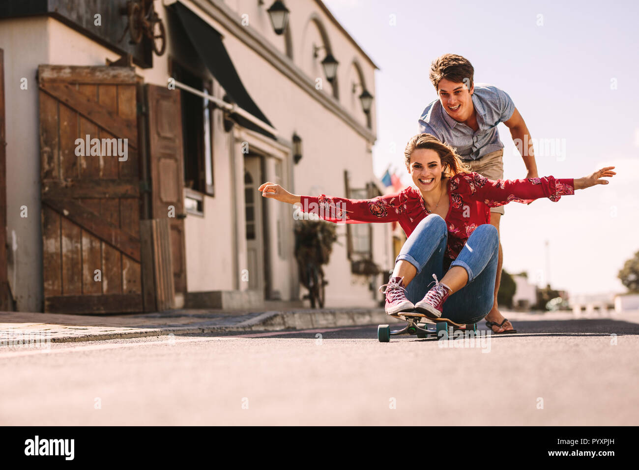 Donna sorridente seduto su uno skateboard lo scorrimento verso il basso una strada vuota. L'uomo aiutando la sua ragazza ride su uno skateboard che spinge da dietro. Foto Stock