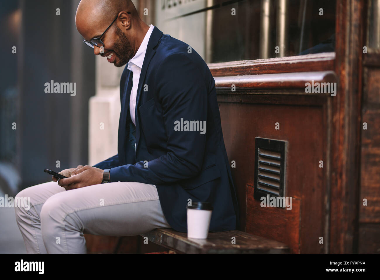 Sorridente giovane seduto su una panchina di legno all'aperto e leggendo messaggi di testo sul suo cellulare. uomo d'affari che usa uno smartphone seduto fuori. Foto Stock