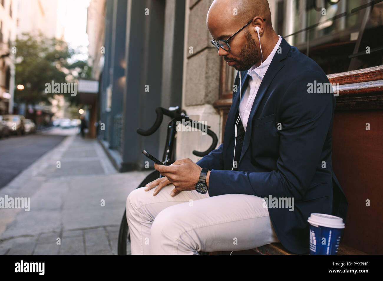 Giovane africano seduto fuori su una panchina usando il telefono cellulare. uomo d'affari che indossa auricolari e ascolta musica dal telefono cellulare. Foto Stock