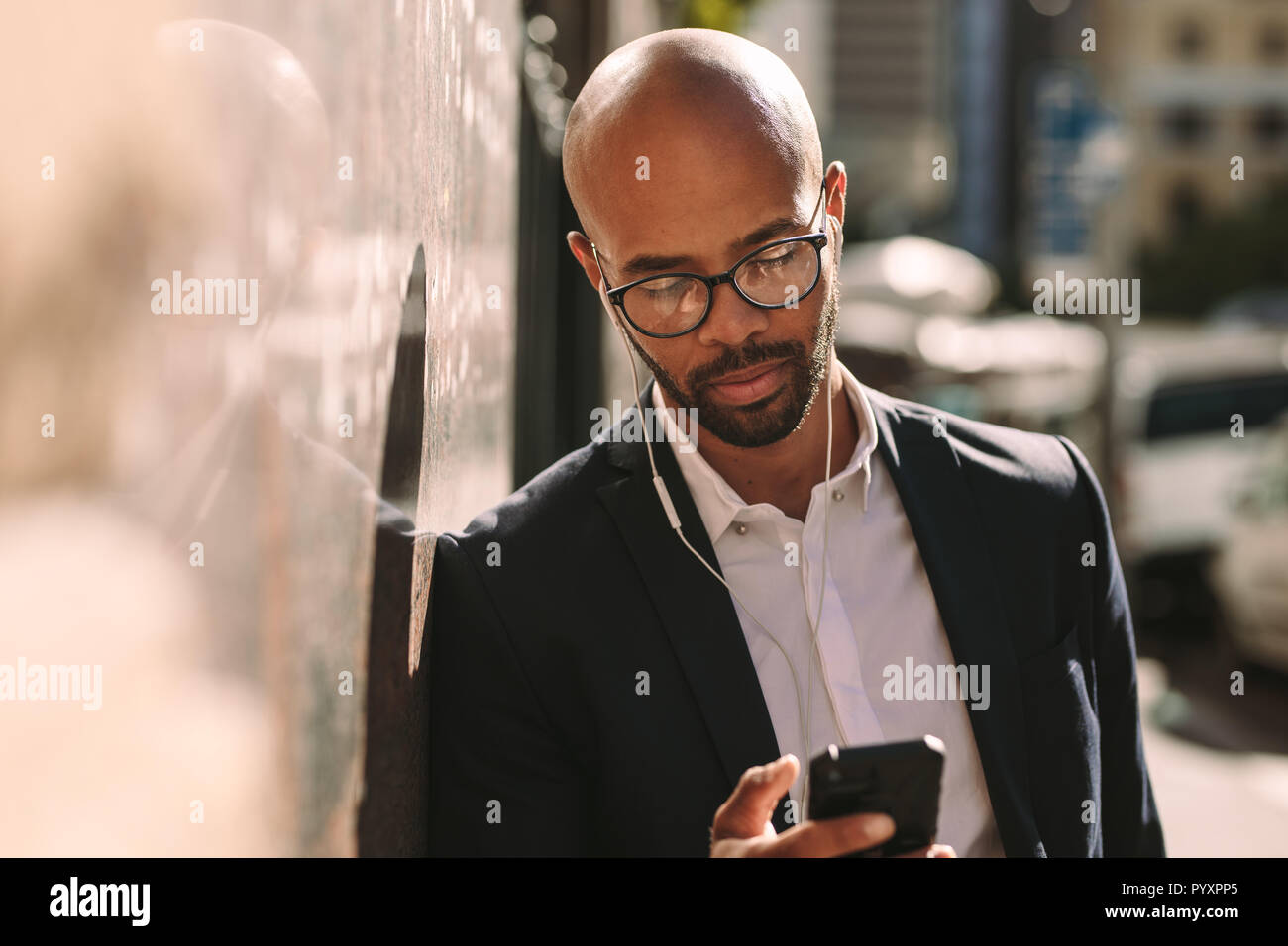 Imprenditore africano il pendolarismo con uno smartphone. Uomo calvo in tuta che indossa gli auricolari tramite telefono cellulare appoggiato alla parete all'esterno. Foto Stock