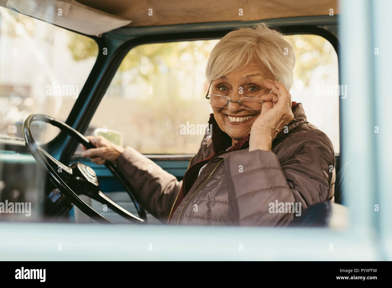 Senior donna seduta nel sedile di guida di auto tenendo gli occhiali in basso per guardare la fotocamera. Il sambuco donna alla guida di una vettura vecchia sul giorno d'inverno. Foto Stock