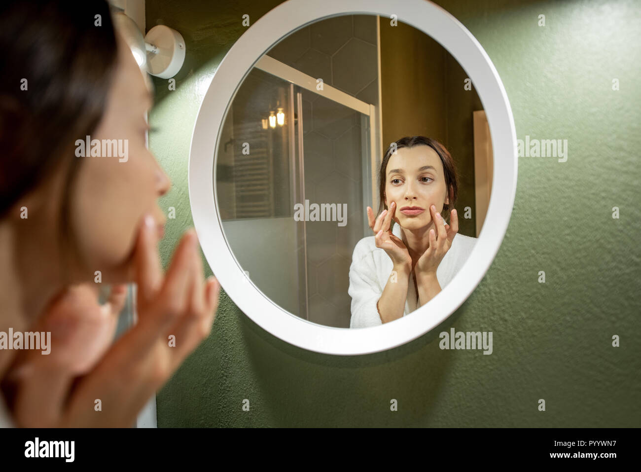 Donna in accappatoio rendendo il massaggio del viso cercando nello specchio rotondo in bagno Foto Stock