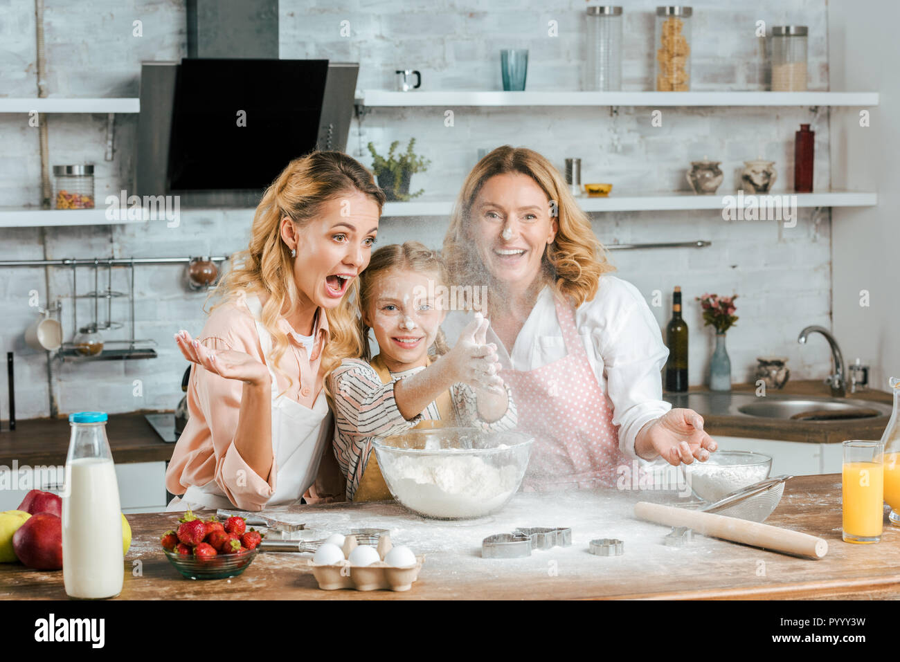 Bambino sorridente battendo le mani con la farina durante la cottura con la madre e la nonna a casa Foto Stock