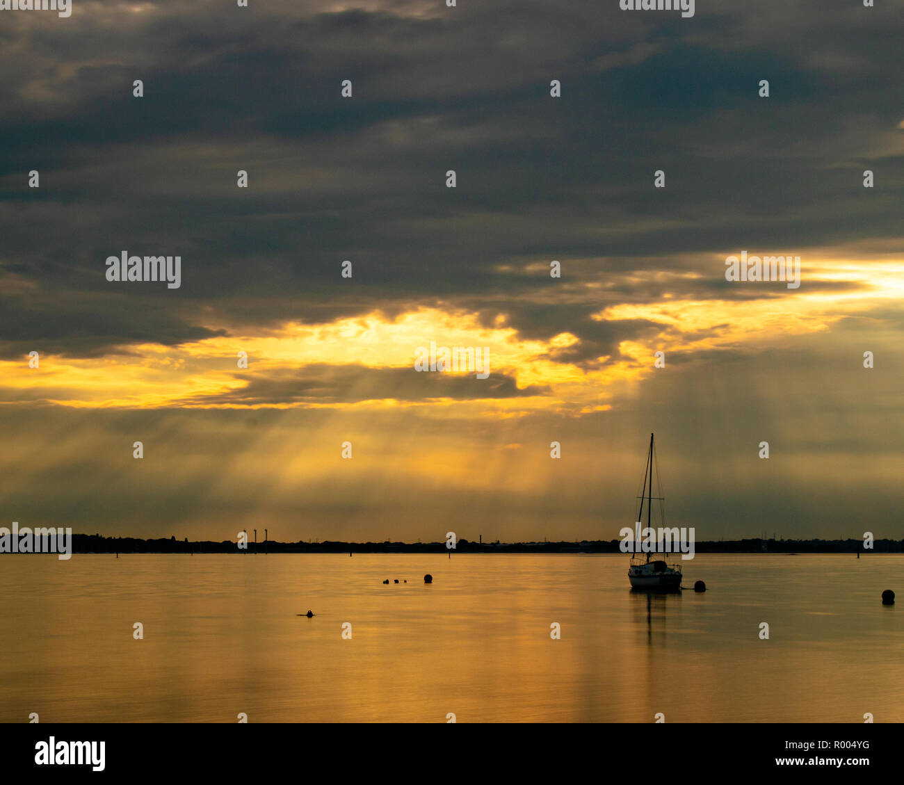 Una sola imbarcazione a vela sul mare calmo al tramonto con i raggi di luce provenienti attraverso le nuvole Foto Stock