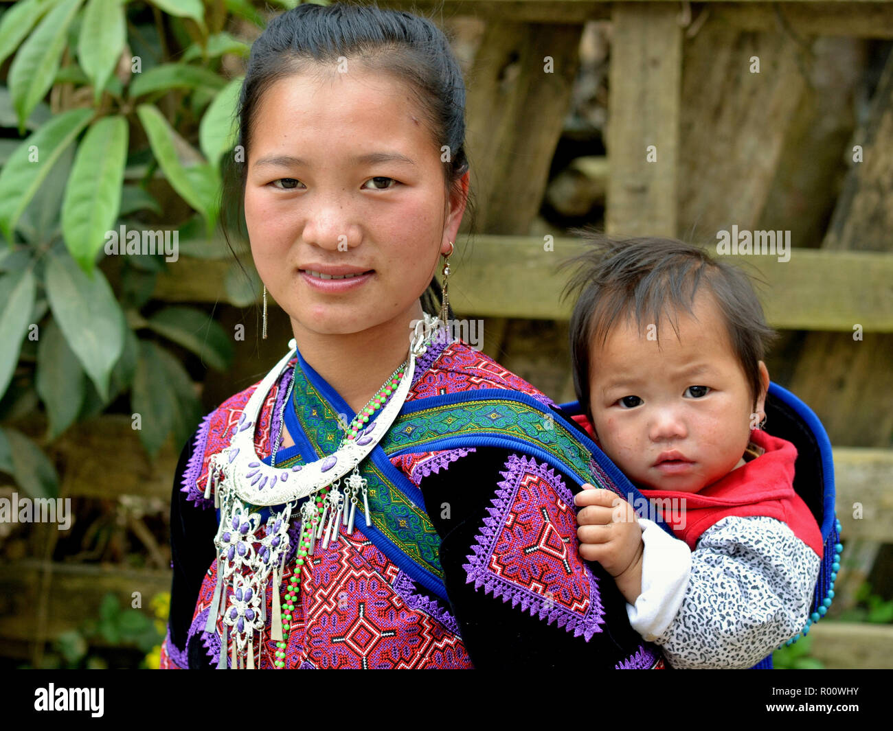 Giovane vietnamita Flower H'mongs hill-tribe donna indossa il suo clan tradizionale di un vestito ricamato e porta il suo bambino il figlio in una fionda bambino sulla schiena. Foto Stock
