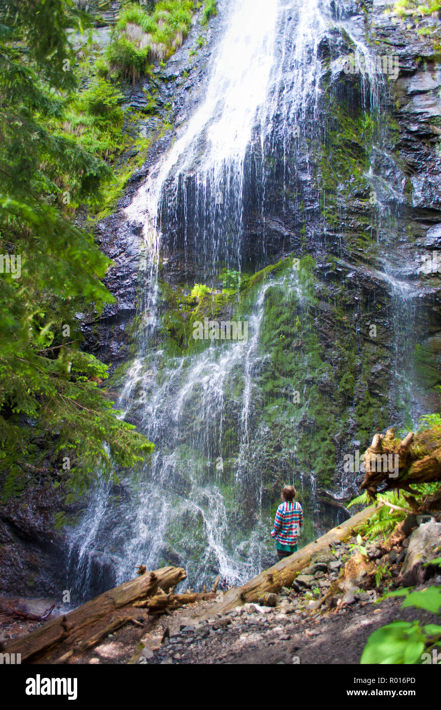 Una giovane ragazza in piedi con la schiena rivolta alla ricerca in remoto in una desolazione Yalynskyi cascata con gocce d'acqua che cadono contro il pendio roccioso. Il verdeggiante pla Foto Stock