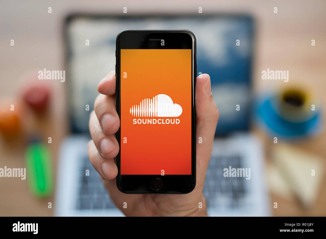 Un uomo guarda al suo iPhone che visualizza il logo Soundcloud, mentre se ne sta seduto alla sua scrivania per computer (solo uso editoriale). Foto Stock