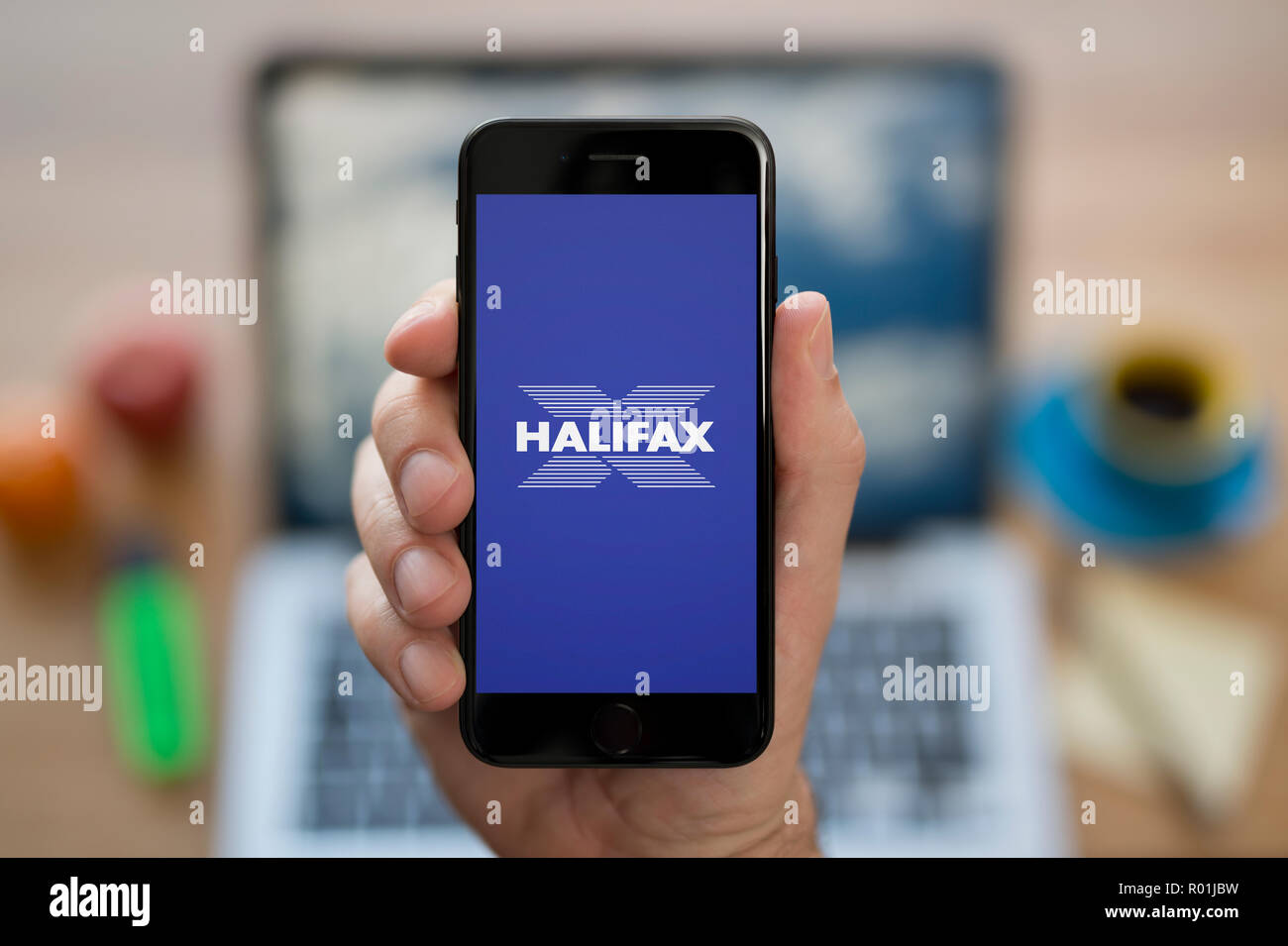 Un uomo guarda al suo iPhone che consente di visualizzare l'Halifax bank logo, mentre se ne sta seduto alla sua scrivania per computer (solo uso editoriale). Foto Stock