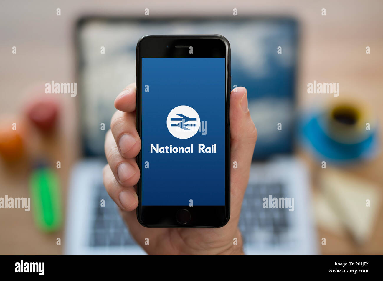Un uomo guarda al suo iPhone che visualizza il National Rail Enquiries logo, mentre se ne sta seduto alla sua scrivania per computer (solo uso editoriale). Foto Stock