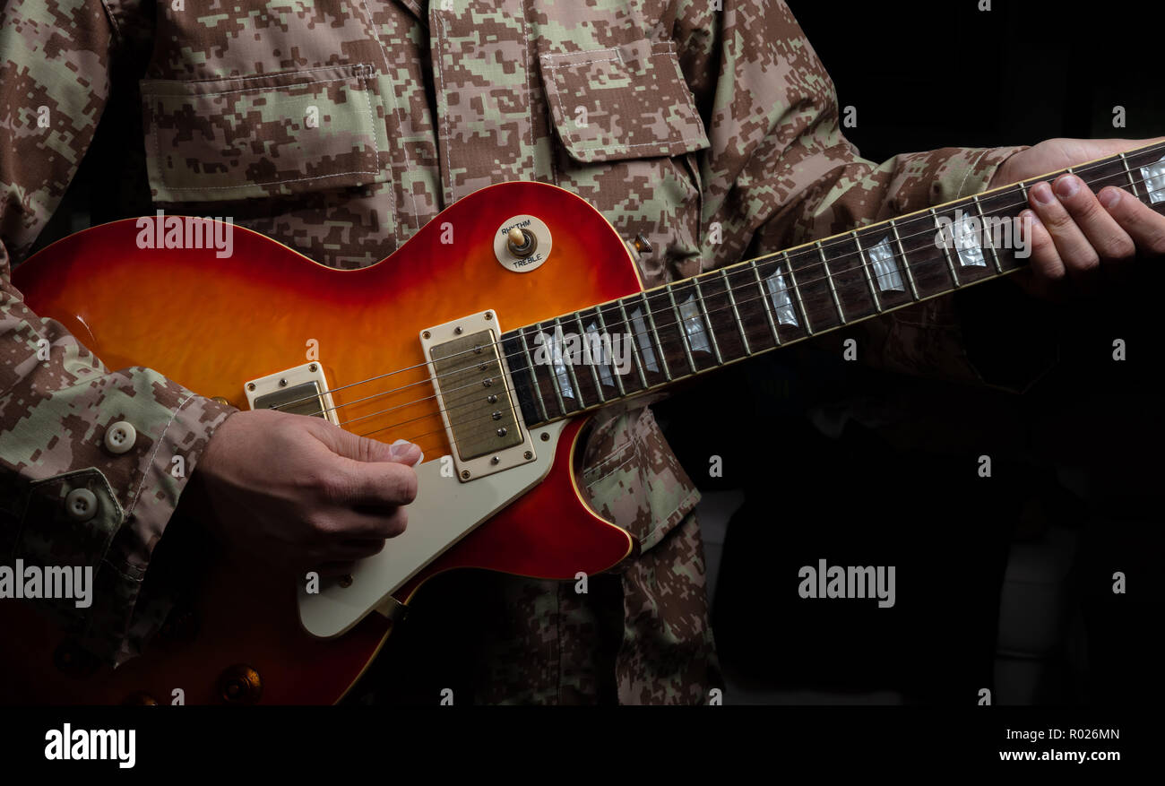 Giovane uomo in uniforme militare suonare la chitarra elettrica, vista ravvicinata, sfondo scuro Foto Stock