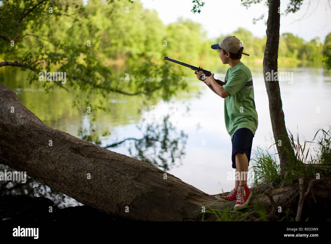 Ragazzo in piedi su un tronco di albero dal lato di un lago con un fucile Foto Stock