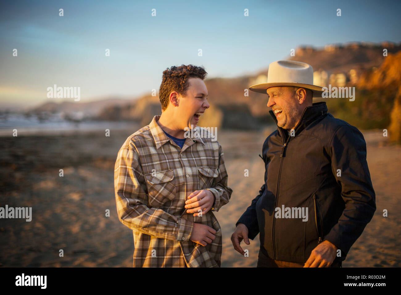 Figlio adolescente e la sua mezza età padre ridere come essi condividono uno scherzo insieme come essi passeggiata sulla spiaggia al tramonto. Foto Stock